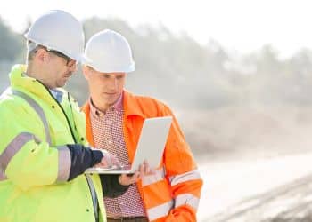 Two safety workers in hard hats and hi-vis reviewing a tablet on a work site