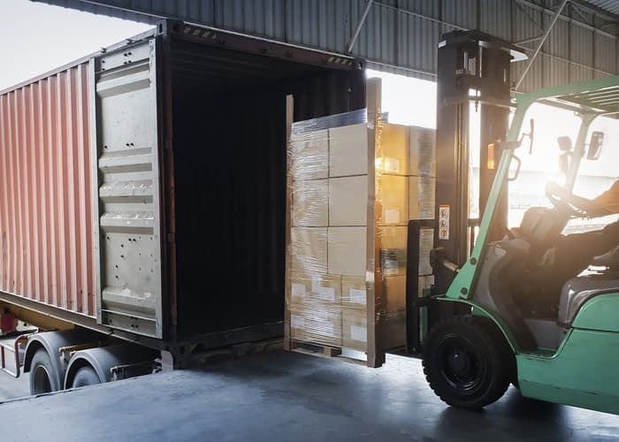 Forklift loading a wrapped pallet of boxes into a shipping container at a loading dock
