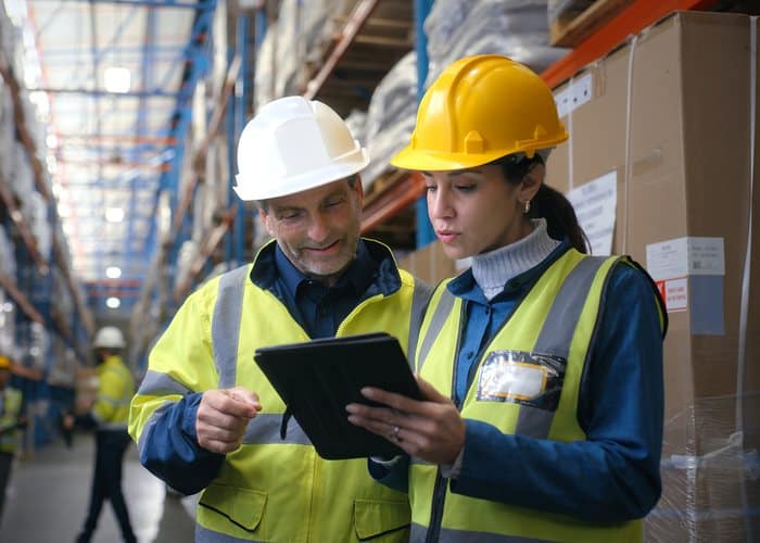 Warehouse supervisor and colleague in hard hats and hi-vis reviewing a tablet between racking aisles